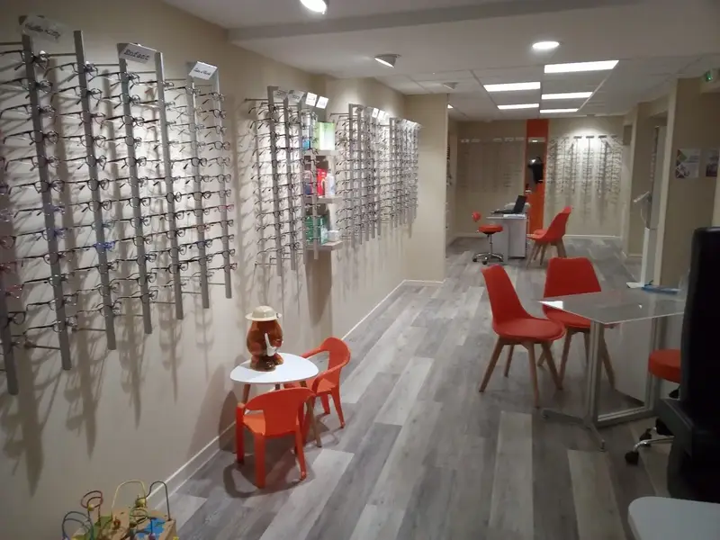 Les présentoirs de montures le long des murs, les chaises orange et la table de conseil